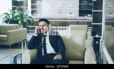 Pan shot di sicuro uomo d'affari parlando telefono cellulare mentre si siede sulla poltrona in hotel di lusso con bagagli vicino a lui. Concetto di viaggi, affari e persone Foto Stock