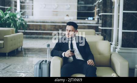 Un'immagine panoramica di un uomo d'affari attento che guarda da parte e usa uno smartphone seduto sulla poltrona in un hotel di lusso con valigia vicino a lui. Concetto di viaggi, affari e persone Foto Stock