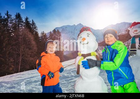 Due felici bambini sorridenti costruiscono e vestono il pupazzo di neve all'esterno Foto Stock