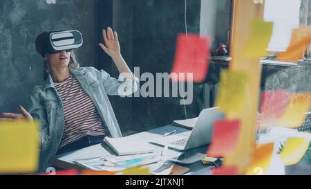 Attraente giovane donna è seduta a tavola e indossando occhiali di realtà virtuale. Sta muovendo le mani e avendo esperienza di realtà virtuale in un moderno ufficio interno Foto Stock