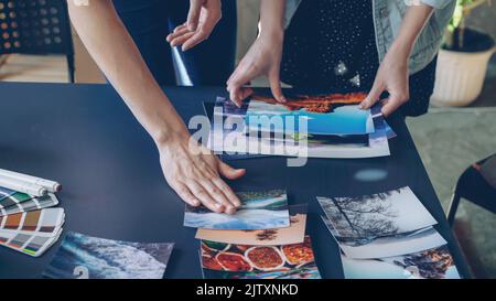 Primo piano delle mani delle donne che collocano le immagini sul tavolo moderno e le dividono in gruppi. Tavolozza di colori, pennarelli e tazza da tè sono sulla scrivania. Concetto di lavoro creativo in corso. Foto Stock