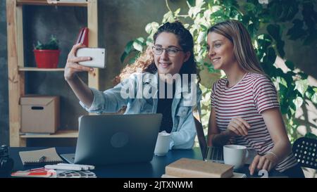 I giovani imprenditori parlano e fanno selfie con lo smartphone mentre si siedono alla scrivania in un ufficio moderno. Stanno posando e ridendo Foto Stock