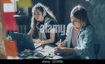 Un team di designer creativi lavora insieme in un ufficio leggero. Allegra signora dai capelli scuri sta ascoltando musica e lavorando con il portatile, e la donna bionda sta disegnando immagini in blocco note Foto Stock