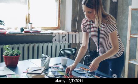 La giovane ragazza bionda fotografa sta facendo flatlay dalle foto colorate sul tavolo e scattandole con la macchina fotografica. Sta visualizzando le immagini vicino alla tavolozza dei colori e ai marcatori sulla scrivania. Foto Stock