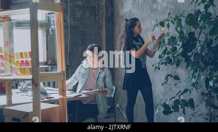 Le amiche e le partner commerciali lavorano insieme in un ufficio moderno. Blonde è seduto a tavola e la raccolta di immagini, brunette è in piedi e innaffiare le piante. Atmosfera informale e accogliente. Foto Stock