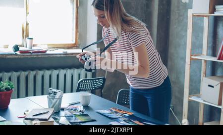 La giovane ragazza bionda fotografa sta creando un piatto disteso dalle foto colorate sul tavolo e scattandole con la fotocamera. Sta visualizzando le immagini vicino alla tavolozza dei colori e ai marcatori sulla scrivania. Foto Stock