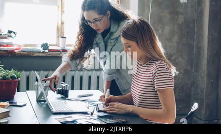La giovane designer bionda sta disegnando immagini nel notebook mentre la sua collega mostra lo schermo del suo notebook e condivide idee. Le donne stanno guardando le foto sul tavolo e controllando la tavolozza dei colori. Foto Stock