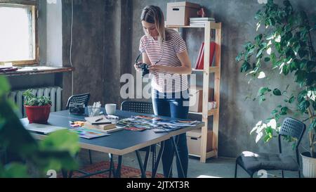 La giovane ragazza creativa sta facendo la disposizione piatta delle foto colorate, quindi scattandole con la macchina fotografica. Donna è in piedi vicino al tavolo moderno in stile loft ufficio e indossare abiti casual. Foto Stock