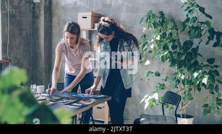 Le giovani imprenditrici stanno mettendo le foto sul tavolo per fare la disposizione piatta e scattare con la macchina fotografica mentre si levano in piedi nell'ufficio leggero moderno. Le donne condividono idee e discutono di design. Foto Stock