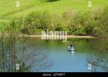 Un uomo che pesca da un boak sul lago artificiale Clatworthy in primavera sulle pendici meridionali delle colline di Brendon, Somerset, Inghilterra. Foto Stock