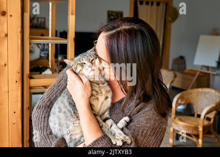 Donna di mezza età che abbraccia carino gatto tabby in scena indoor. Relazioni uomo-animale. Animale domestico divertente. Animali domestici senza casa. Foto Stock