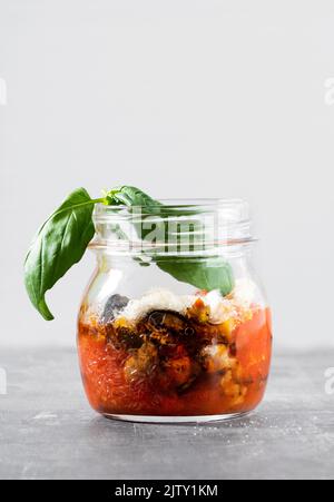 Parmigiana di melanzane - melanzana al forno con formaggio, pomodori e spezie, servita in un vasetto di vetro con basilico fresco Foto Stock