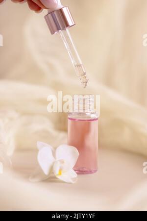 Gocce di siero da una pipetta in un flacone cosmetico aperto vicino a fiori di orchidee bianche su primo piano beige chiaro. Prodotti di bellezza per la cura della pelle, essenze o siero. E Foto Stock