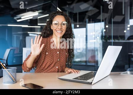 Ritratto di una giovane donna di successo in ufficio moderno utilizzando un computer portatile, donna ispanica con capelli ricci sorridendo e guardando la fotocamera, tenendo la mano in su nel gesto di saluto. Foto Stock