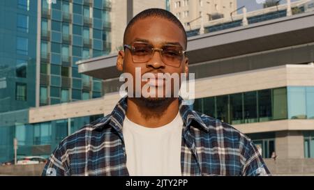 Ritratto di uomo in città african american ragazzo giovane studente uomo in occhiali da sole di stile indossa plaid camicia sguardi posa sognando pensare contempla Foto Stock