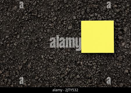 Giallo nota carta quadrato su suolo nero struttura terra primo piano. Giallo sticky note carta bianca terreno piano terra organica struttura. Mockup su suolo di terra Foto Stock