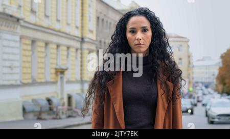 Serio solo sicuro potente giovane ragazza bruna con capelli lunghi ricci business donna con espressione arrabbiata si trova sulla strada in città guardando Foto Stock