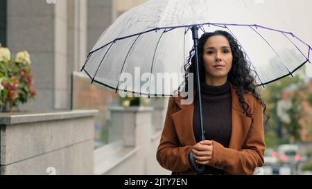 Calma seria femmina turista giovane bella donna sognante ispirata ragazza signora con lunghi capelli ricci con ombrello trasparente passeggiate in città piovosa Foto Stock