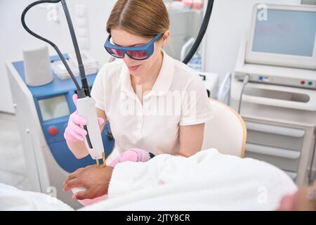 Dermatologo in occhiali di sicurezza che esegue la rimozione dei peli sulla pelle del paziente Foto Stock