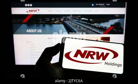 Persona che detiene uno smartphone con il logo della società mineraria australiana NRW Holdings Limited sullo schermo di fronte al sito Web. Messa a fuoco sul display del telefono. Foto Stock