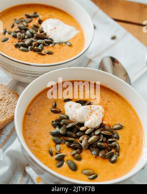 Zuppa di paprika cremosa, zuppa di crema con panna acida e semi di zucca nel recipiente sul tavolo con pane fresco e cucchiai Foto Stock