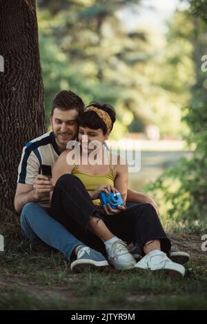 Bella coppia che guarda la foto mentre si siede sull'erba vicino all'albero nel parco Foto Stock