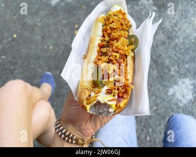 L'uomo tiene in mano un cane caldo con cipolle fritte e peperoni. Messa a fuoco selettiva. Cibo di strada Foto Stock