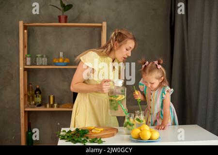 la mamma versa la limonata in bicchieri, e la figlia mette i tubetti in un bicchiere. bevete il succo di limone fresco in cucina Foto Stock