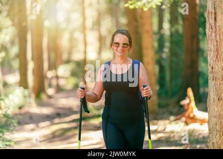 Donna con gli occhiali sorridenti e camminando attraverso la foresta con i pali escursionistici. I raggi del sole si rompono attraverso gli alberi Foto Stock