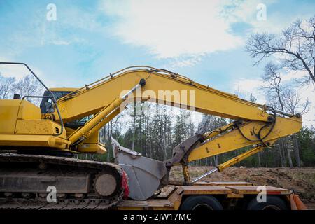 Braccio e benna di una pala gommata SSL gialla su un rimorchio a pianale, in cantiere, con luce notturna e alberi sullo sfondo. Foto Stock