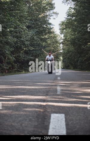 Una giovane coppia felice guida una moto su una strada asfaltata nella foresta, la libertà e la velocità Foto Stock
