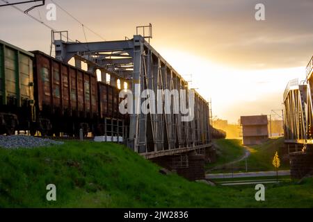 Un ponte ferroviario sul quale un treno con vagoni si muove sullo sfondo di un sole luminoso Foto Stock