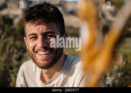 Felice giovane maschio bearded in t shirt bianca con orecchini sorridente e guardando la macchina fotografica mentre si passa il tempo in campagna in estate Foto Stock