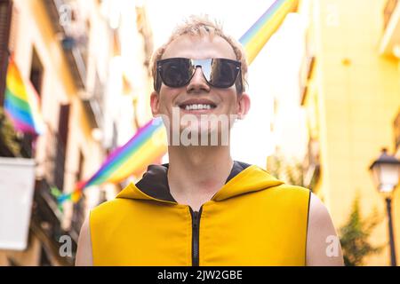 Da sotto felice non binary maschio in abiti eleganti sorridendo in occhiali da sole su sfondo sfocato di case decorate con bandiere LGBT Foto Stock