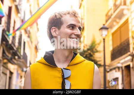 Da sotto felice non binary maschio in abiti eleganti sorridendo e guardando lontano su sfondo sfocato di case decorate con bandiere LGBT Foto Stock
