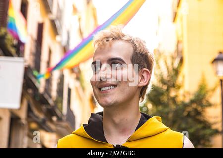 Da sotto felice non binary maschio in abiti eleganti sorridendo e guardando lontano su sfondo sfocato di case decorate con bandiere LGBT Foto Stock