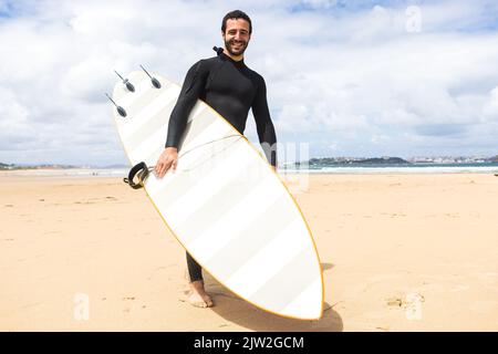 Corpo pieno di giovane giovane giovane surfista ispanico con capelli scuri e barba in muta nera sorridente mentre si è in piedi con tavola da surf gialla sulla spiaggia sabbiosa Foto Stock