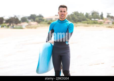 Corpo pieno di giovane giovane giovane giovane surfista ispanico con capelli scuri e in muta sorridente mentre si trova in piedi con tavola da surf blu sulla spiaggia di sabbia Foto Stock