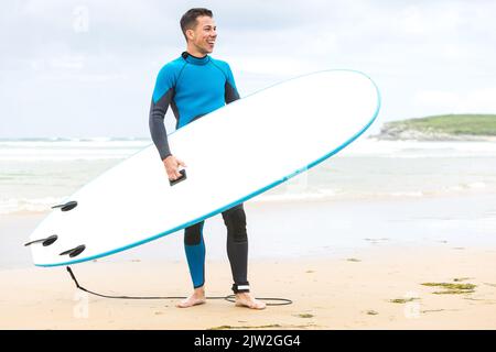 Corpo pieno di giovane giovane giovane giovane surfista ispanico con capelli scuri e in muta sorridente mentre si trova in piedi con tavola da surf bianca sulla spiaggia di sabbia Foto Stock