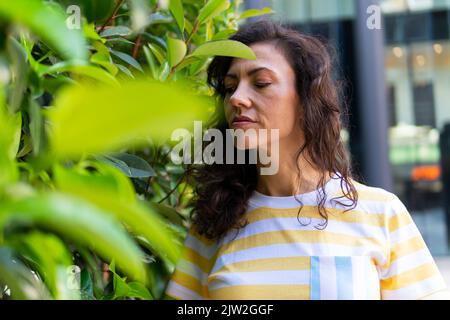 Donna penosa con occhi chiusi in piedi vicino a cespuglio con verde fogliame sulla strada vicino edificio il giorno d'estate in città Foto Stock