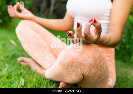 Crop femmina in abbigliamento sportivo meditando con le mani mudra mentre si siede sul prato durante l'allenamento yoga nel parco Foto Stock