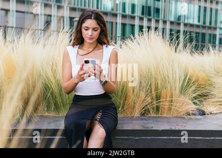 Giovane donna seria con lunghi capelli castani in elegante top e gonna seduta su panca di pietra vicino erba secca e messaggi sullo smartphone nel parco cittadino Foto Stock