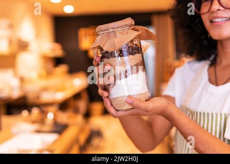 Giovane donna etnica felice tagliata irriconoscibile in uniforme e bicchieri in vetro con frutta a guscio e farine diverse mentre si lavora in zero rifiuti groc Foto Stock