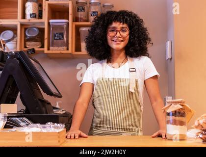 Giovane donna afro-americana sorridente in grembiule e occhiali guardando la macchina fotografica in piedi vicino al moderno registratore di cassa durante il lavoro in zero rifiuti gr Foto Stock