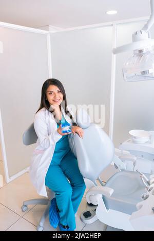 Contenuto giovane dentista ispanica femminile con lunghi capelli scuri in uniforme medica sorridente e guardando la macchina fotografica mentre si siede vicino a sedia dentale in clinica Foto Stock