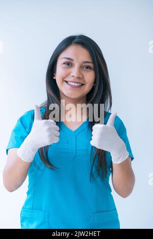 Felice giovane medico etnico con lunghi capelli scuri in scrub blu e guanti in lattice sorridente e mostrando pollici verso l'alto gesto mentre si guarda la macchina fotografica Foto Stock