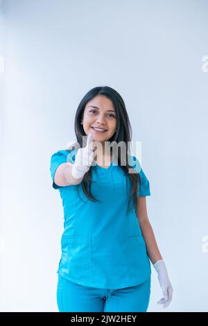 Felice giovane medico etnico con lunghi capelli scuri in scrub blu e guanti in lattice sorridente e mostrando pollici verso l'alto gesto mentre si guarda la macchina fotografica Foto Stock