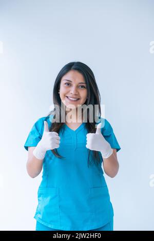 Felice giovane medico etnico con lunghi capelli scuri in scrub blu e guanti in lattice sorridente e mostrando pollici verso l'alto gesto mentre si guarda la macchina fotografica Foto Stock