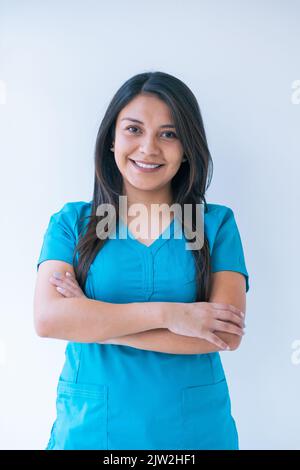 Felice giovane medico etnico femminile con lunghi capelli scuri in scrub blu con braccia incrociate mentre si guarda la macchina fotografica in clinica Foto Stock
