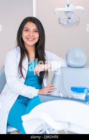 Contenuto giovane dentista ispanica femminile con lunghi capelli scuri in uniforme medica sorridente e guardando la macchina fotografica mentre si siede vicino a sedia dentale in clinica Foto Stock